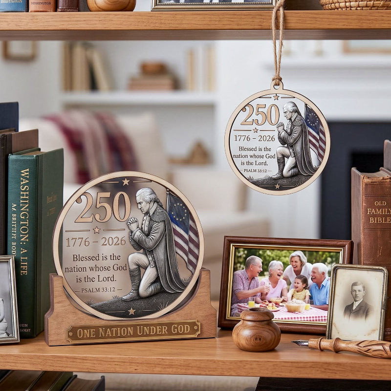 A decorative round hanging ornament and matching plaque on a wooden base, both featuring a kneeling figure, the year 250 1776–2026, and the text 'Blessed is the nation whose God is the Lord.' The ornaments are styled on a wooden shelf with books, a family photo frame, and small decor items in a home interior setting.