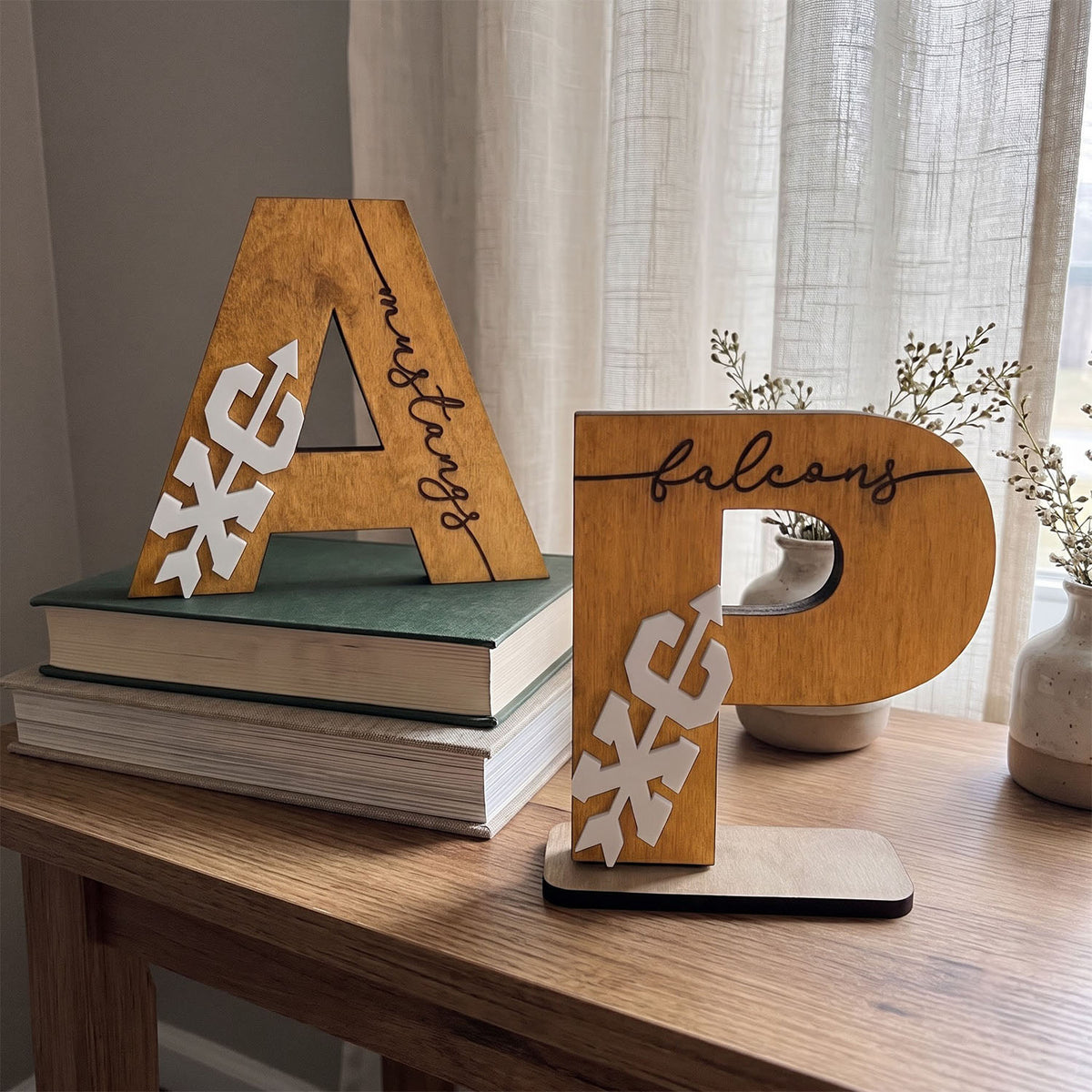 Two wooden decorative letters, 'A' and 'P', displayed on a wooden table. The letter 'A' is stacked on two books and has 'my family' written on it, while the letter 'P' stands on a small base and has 'falcons' written on it.