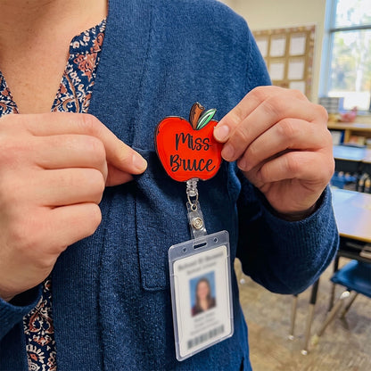 A person in a blue cardigan holds a red apple-shaped pin that says 'Miss Bruce' in front of their chest, with a clear ID badge attached below on a class ID lanyard. The background shows a blurred classroom setting.