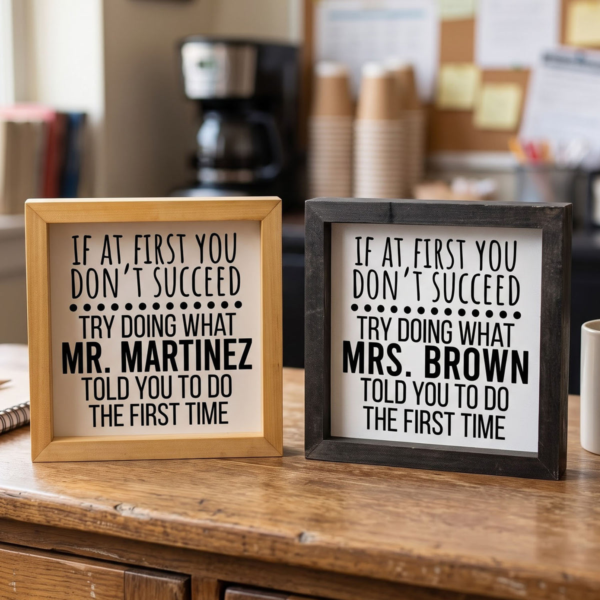 Two humorous framed signs on a wooden table in a cafe setting. One has a light wood frame and the other has a dark charcoal frame, both with white backgrounds and black text reading 'IF AT FIRST YOU DON'T SUCCEED'.