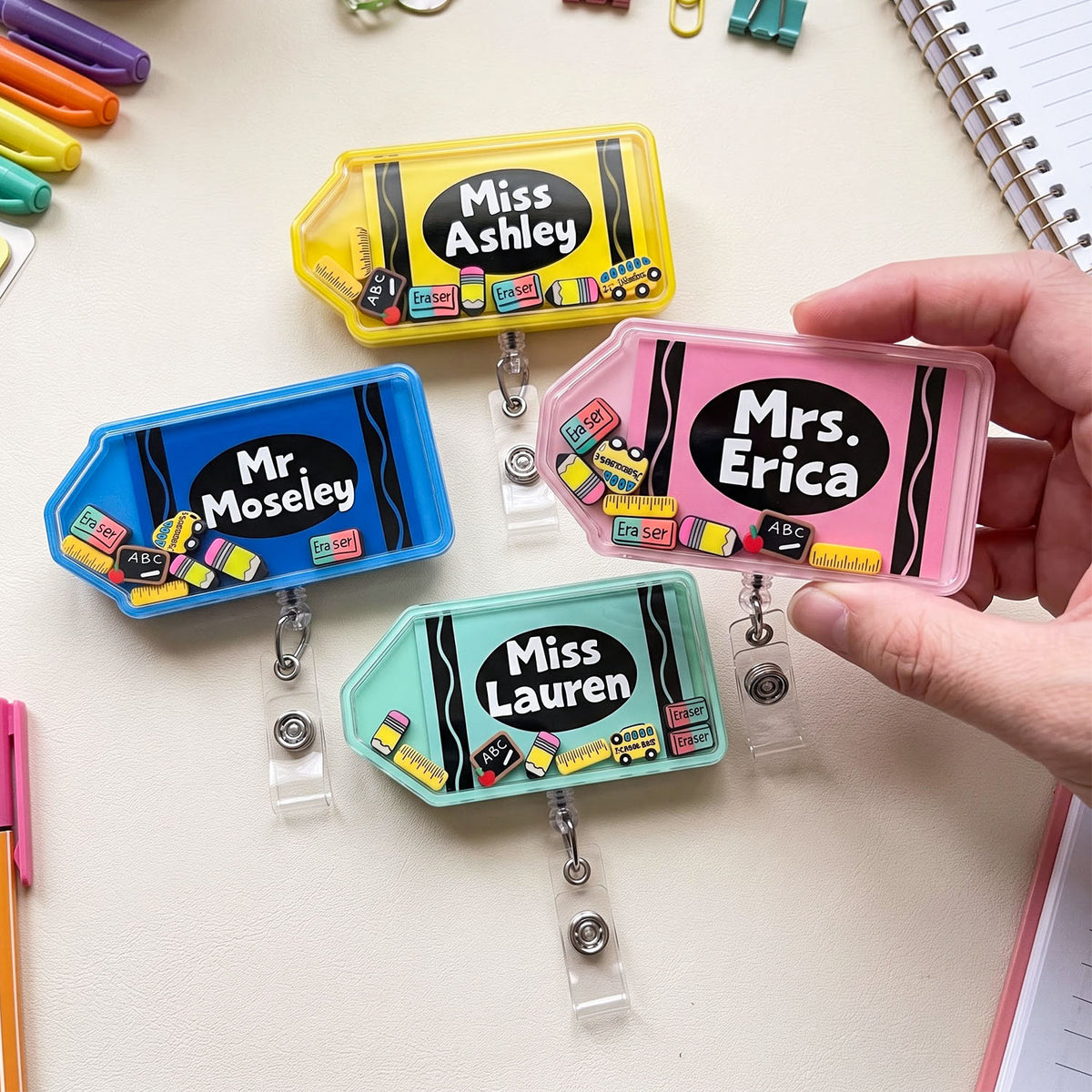 Four colorful retractable badge reel nameplates are arranged on a light desk surface, with one held by a hand. The nameplates come in yellow, blue, pink, and mint green, each with a black oval label area containing white text and small multicolor desk-themed illustrations.