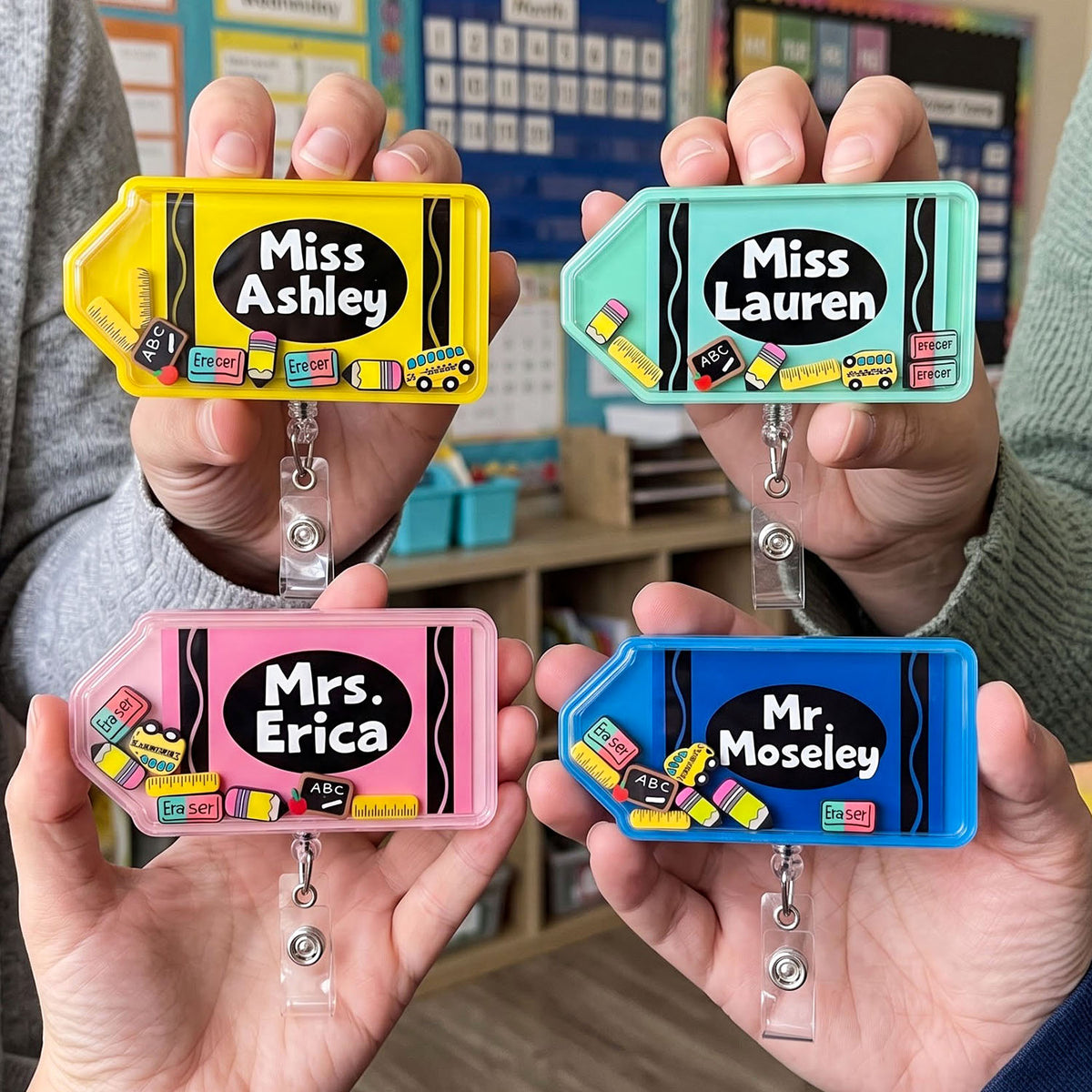 Four colorful retractable badge reel holders are shown being held by hands in a classroom setting. The badges are yellow, mint green, pink, and blue, each featuring a black oval with white text reading 'Miss Ashley', 'Miss Lauren', 'Mrs.