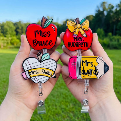 Two hands holding personalized school-themed retractable badge reels outdoors. The left reel is shaped like a red apple with the text 'Miss Bruce' and the right reel is also red with 'Mrs.