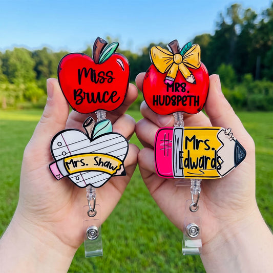 Two hands holding personalized school-themed retractable badge reels outdoors. The left reel is shaped like a red apple with the text 'Miss Bruce' and the right reel is also red with 'Mrs.