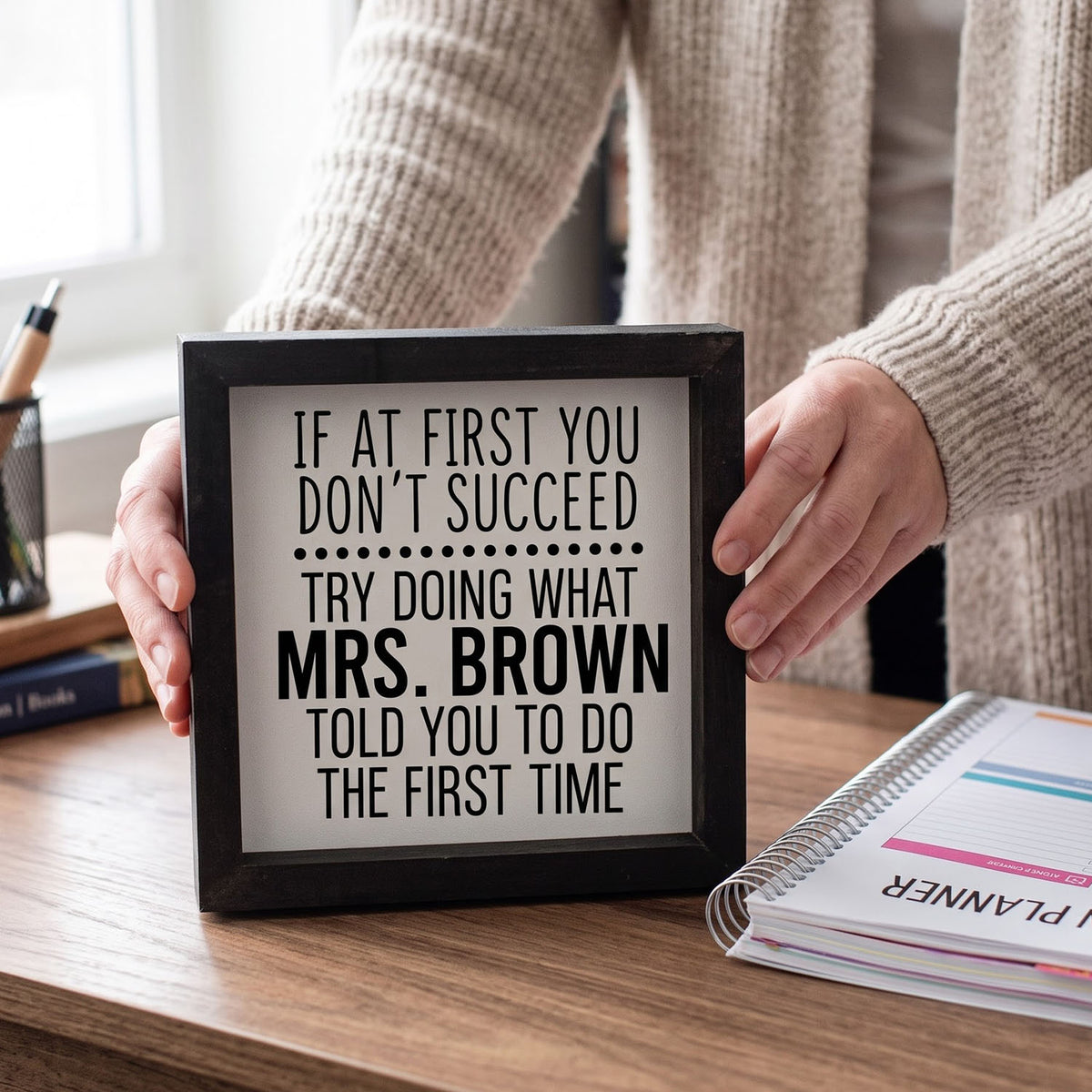A person in a beige knit sweater holds a black-framed sign with a humorous quote about Mrs. Brown on a wooden desk next to a planner.
