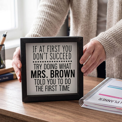 A person in a beige knit sweater holds a black-framed sign with a humorous quote about Mrs. Brown on a wooden desk next to a planner.