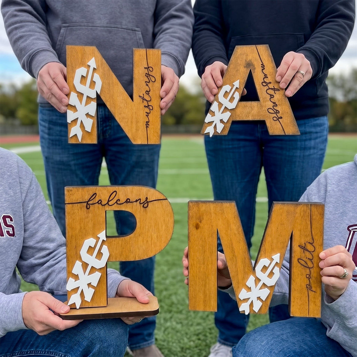 Four people standing on a grassy field hold large wooden letters spelling out a custom name or initials, with white cutout snowflake shapes layered inside some letters. The letters include N, A, P, M, and smaller letters forming words like 'balcony' and 'maliti'.