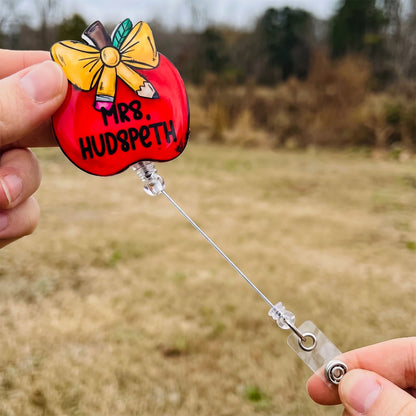 A hand holds a red apple-shaped retractable badge reel with a yellow bow and the text 'MR. HUDSPETH' in front of an outdoor grassy field.