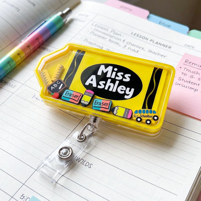 A bright yellow retractable badge reel pencil case with a clear plastic cover, featuring a black oval with the text 'Miss Ashley' and small school-themed illustrations. It is shown lying on a lined notebook page with a pen, sticky notes, and colorful tabs around it.