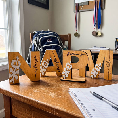 Wooden decorative letters spelling 'NAR' arranged on a wooden desk in a home office setting, with a blue backpack, medals on a wall, and an open notebook with a pen in the foreground.