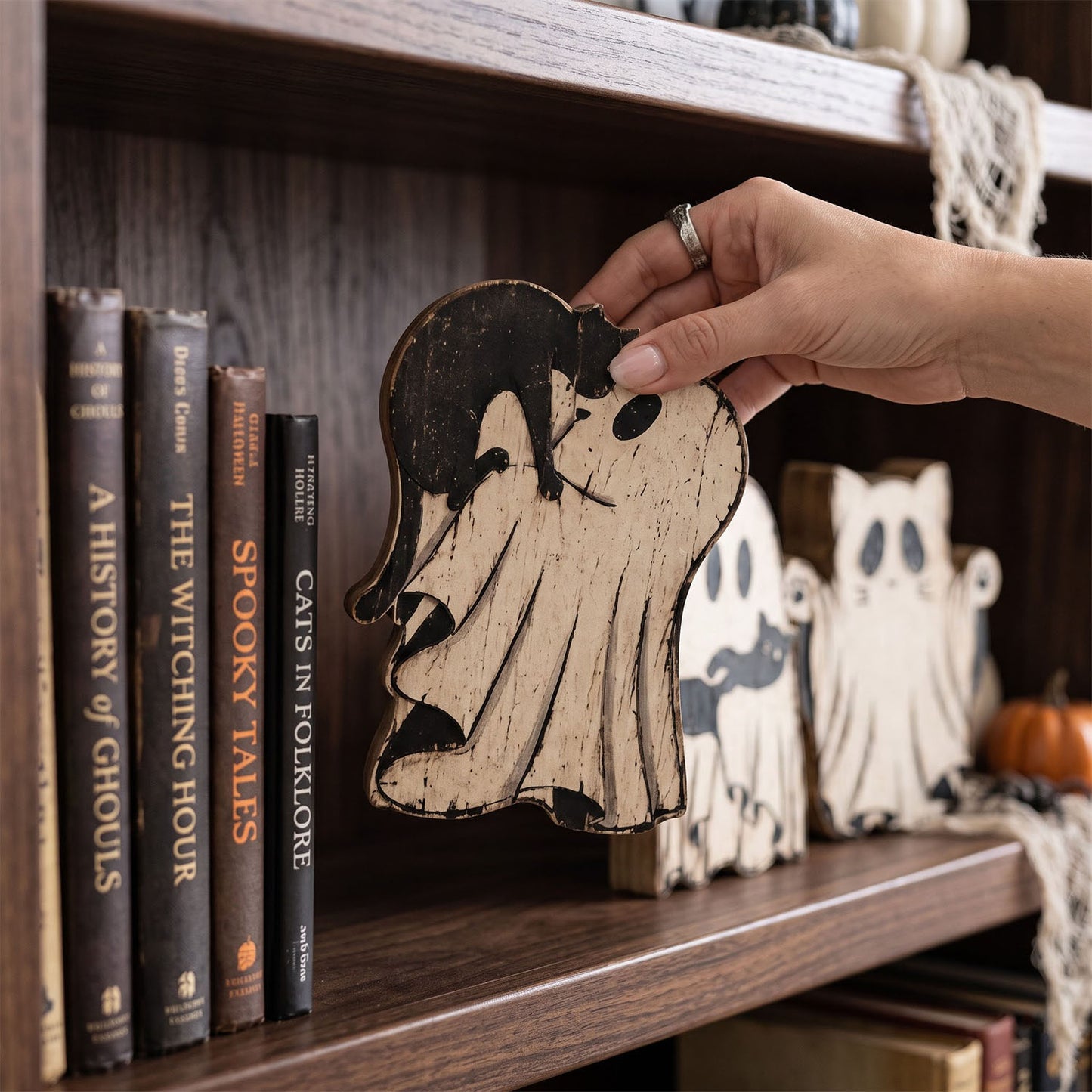 A hand holds a rustic wooden Halloween ghost ornament with a distressed black and natural wood finish, set against a cozy bookshelf background filled with books and seasonal decor.