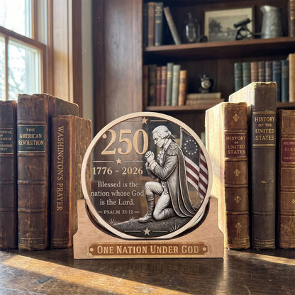 A round wooden plaque featuring an engraved depiction of George Washington kneeling with the American flag, flanked by the numbers 250 and 1776-2026, with the text 'Blessed is the nation whose God is the Lord' and 'ONE NATION UNDER GOD' at the bottom. The plaque is displayed on a wooden table between vintage books in a library setting.