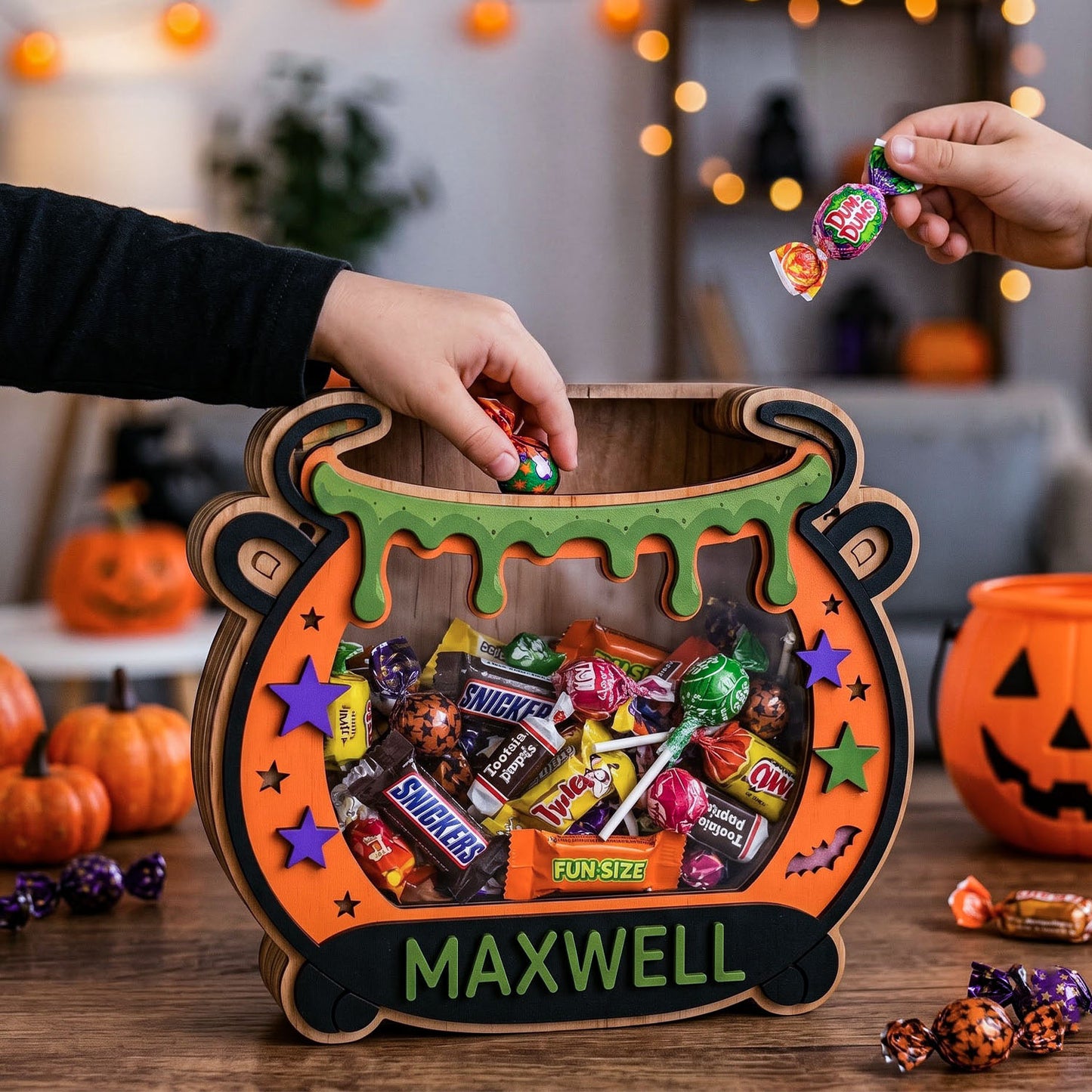 A Halloween-themed cauldron-shaped candy bucket filled with assorted chocolates and treats, shown on a table with hands placing and taking candies. The bucket has green dripping trim, purple and green stars, a bat detail, and the name 'MAXWELL' on the front.
