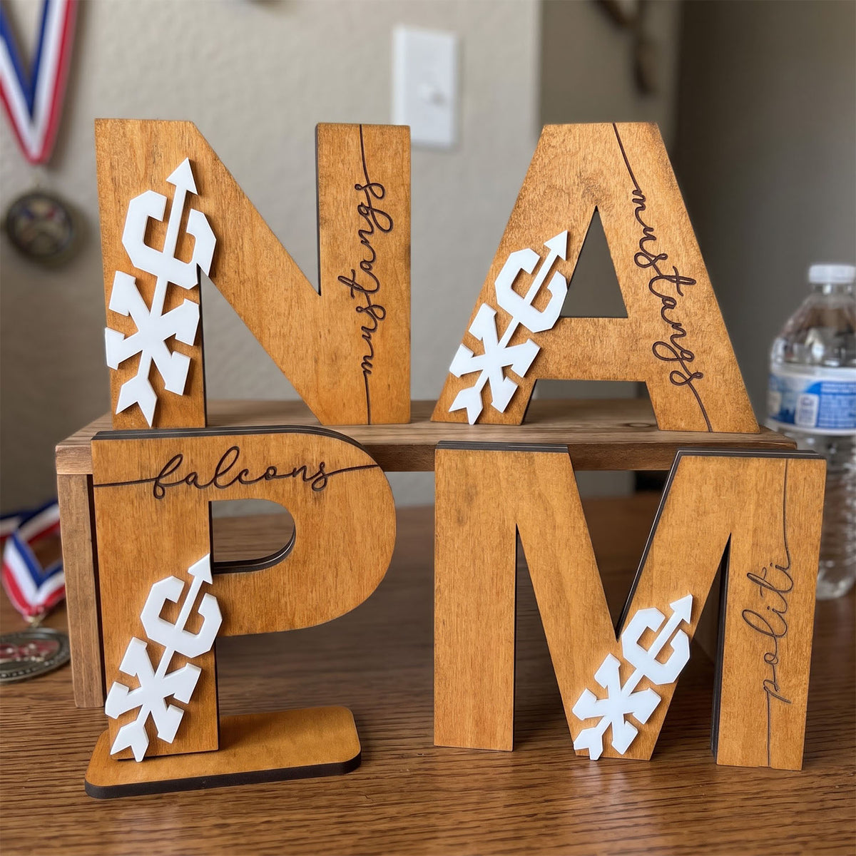 Wooden decorative letters spelling NAFAFLM with white arrow and snowflake cutouts, displayed on a wooden stand on a tabletop.