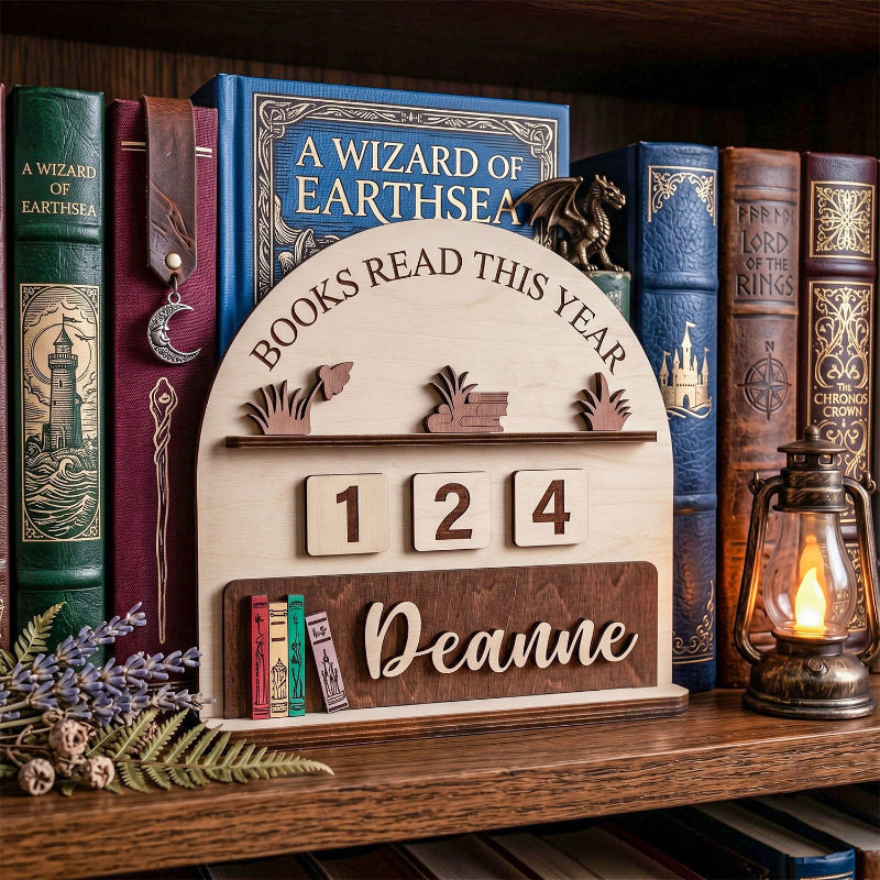 A wooden bookshelf plaque displayed on a wooden bookshelf, surrounded by vintage-style books and a lit lantern. The plaque is light natural wood with a darker wood base, featuring the text 'BOOKS READ THIS YEAR', numbers '1 2 4', and the name 'Deanne'.