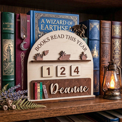 A wooden bookshelf plaque displayed on a wooden bookshelf, surrounded by vintage-style books and a lit lantern. The plaque is light natural wood with a darker wood base, featuring the text 'BOOKS READ THIS YEAR', numbers '1 2 4', and the name 'Deanne'.