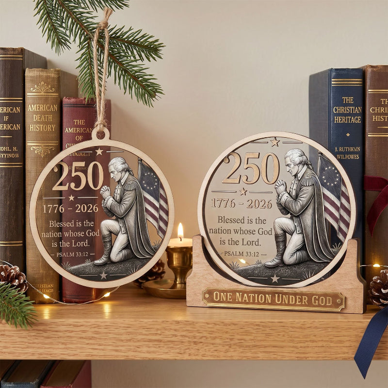 Two round wooden Christmas ornaments displayed on a wooden shelf, one hanging and one resting on a small wooden stand. The ornaments feature a silver-toned relief of a kneeling patriotic figure with an American flag background and the text '250 1776-2026 Blessed is the nation whose God is the Lord.