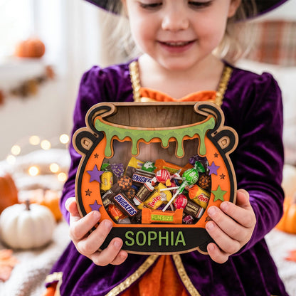 A child in a purple witch costume holds a wooden cauldron-shaped candy box filled with assorted wrapped chocolates and gummy candies. The front of the box is personalized with the name 'SOPHIA' in green letters on a black band, with orange, purple, green, and brown details.