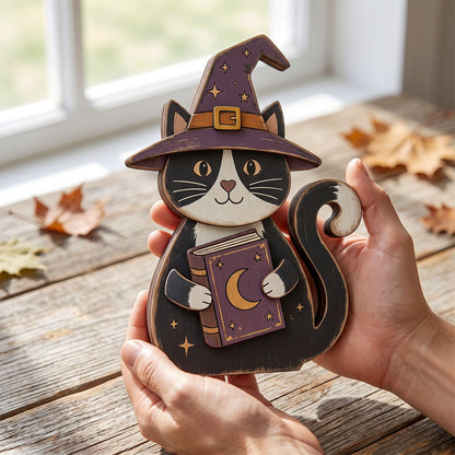 A wooden cat-shaped decorative piece wearing a witch hat and holding a book, being held by two hands over a rustic wooden table with scattered autumn leaves near a window.