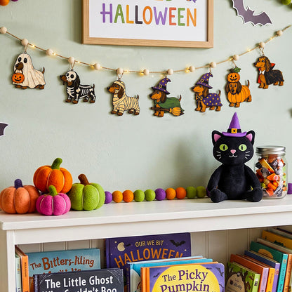 A styled Halloween shelf display featuring a felt garland of cute dog characters, a plush black cat, colorful felt pumpkins, and a black cat hat, set against a light green wall with a framed 'HALLOWEEN' sign.
