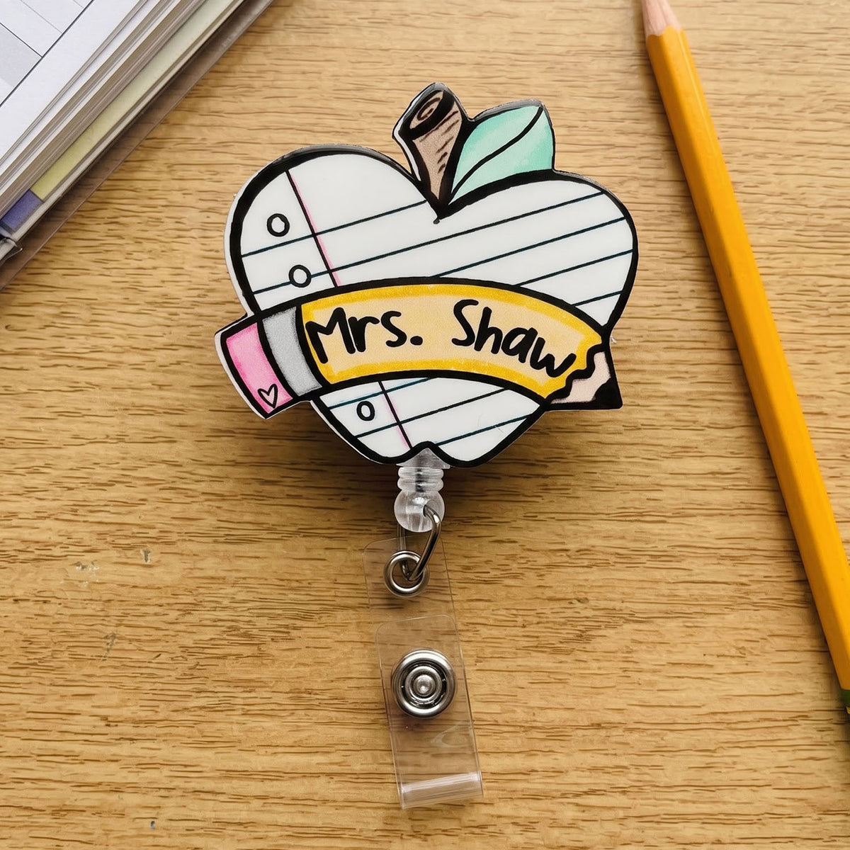 A novelty retractable badge reel featuring a cartoon apple with lined notebook paper, a brown stem, green leaves, a pink eraser, and a yellow banner reading 'Mrs. Shaw'.