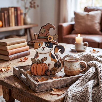 A decorative cat figurine wearing a witch hat and holding a pumpkin sits on a rustic wooden tray on a table, styled with a ceramic mug, a knitted throw blanket, scattered autumn leaves, and a softly lit living room in the background.