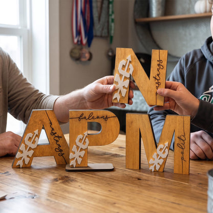 Four wooden letter blocks spelling out 'APM' and 'N' are arranged on a wooden table, with parts of two people holding and assembling the letters visible in a home setting.