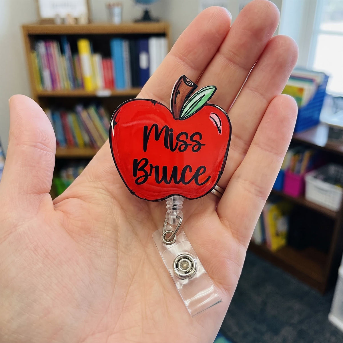 A hand holding a red apple-shaped retractable badge reel with the text 'Miss Bruce' in black script, set against a blurred classroom background with bookshelves.