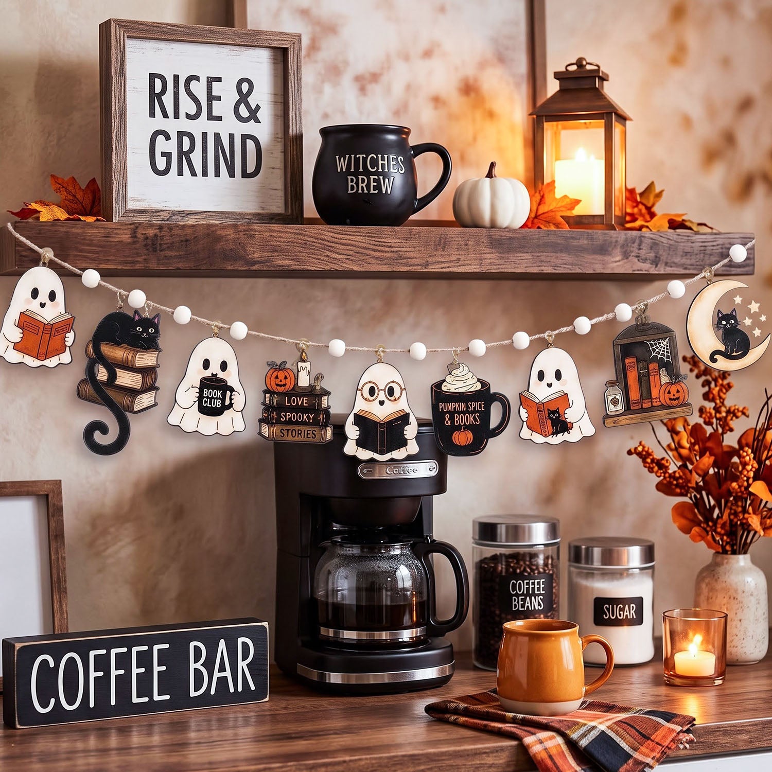 A styled autumn coffee bar shelf display with a wooden 'RISE & GRIND' sign, a black 'WITCHES BREW' mug, a lantern, fall leaves, a pumpkin, and a string banner featuring ghost, owl, pumpkin, and tombstone shapes. Below the shelf is a coffee maker, a coffee bean jar, a sugar jar, an orange mug, and a plaid napkin against a warm beige wall.