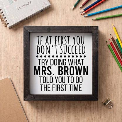 A framed typographic sign with the text “IF AT FIRST YOU DON’T SUCCEED TRY DOING WHAT MRS. BROWN TOLD YOU TO DO THE FIRST TIME” in black lettering on a white background, set in a dark wood frame on a light wood desk with colored pencils and stationery around it.
