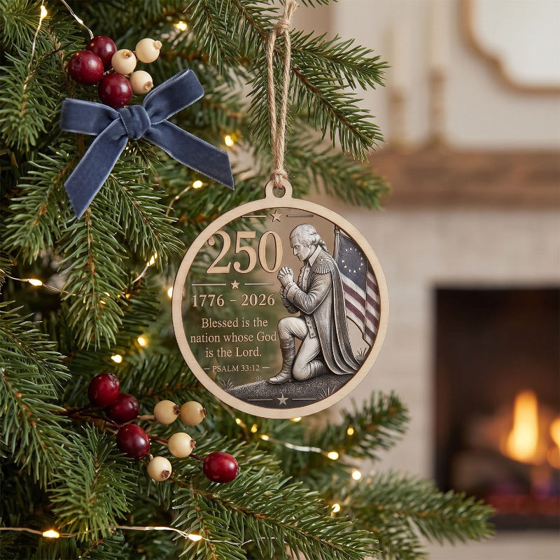 A round wooden Christmas ornament hanging from a tree branch, featuring a relief design of a kneeling praying man, the American flag, and the text '250 1776–2026 Blessed is the nation whose God is the Lord. Psalm 33:12.