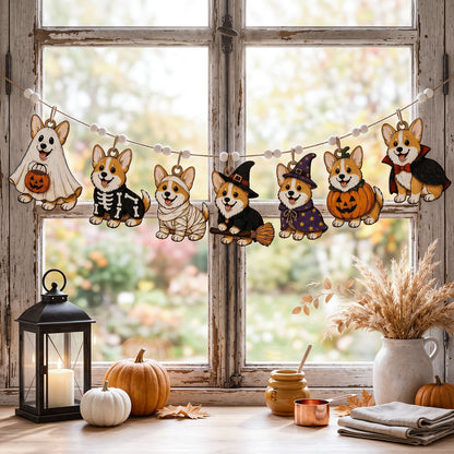 A Halloween-themed decorative garland featuring cartoon corgi dogs in ghost, skeleton, mummy, witch, pumpkin, and vampire costumes, hanging across a rustic wooden window. The scene is styled with autumn decor including pumpkins, a lantern, a ceramic vase with dried grass, and folded fabric on a tabletop.