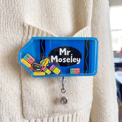 A blue retractable badge reel clip shaped like a crayon, pinned to a cream ribbed sweater. The clip features a black oval with the text 'Mr Moseley' in white, surrounded by colorful toy eraser and chalk illustrations.