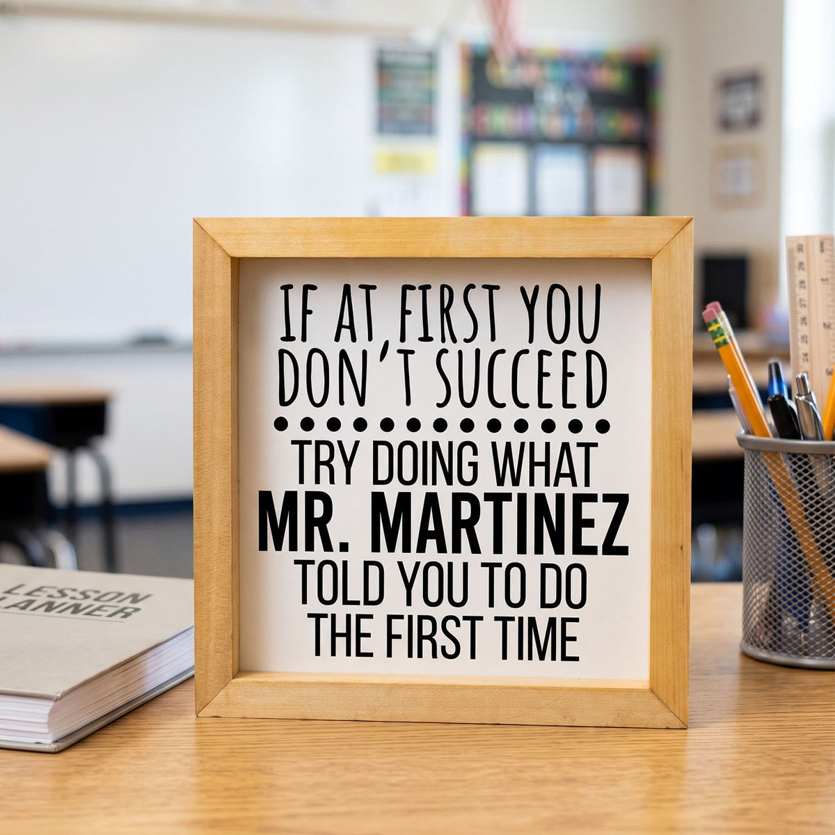 A framed typographic sign on a wooden desk in a classroom setting, reading: 'IF AT FIRST YOU DON'T SUCCEED TRY DOING WHAT MR. MARTINEZ TOLD YOU TO DO THE FIRST TIME.
