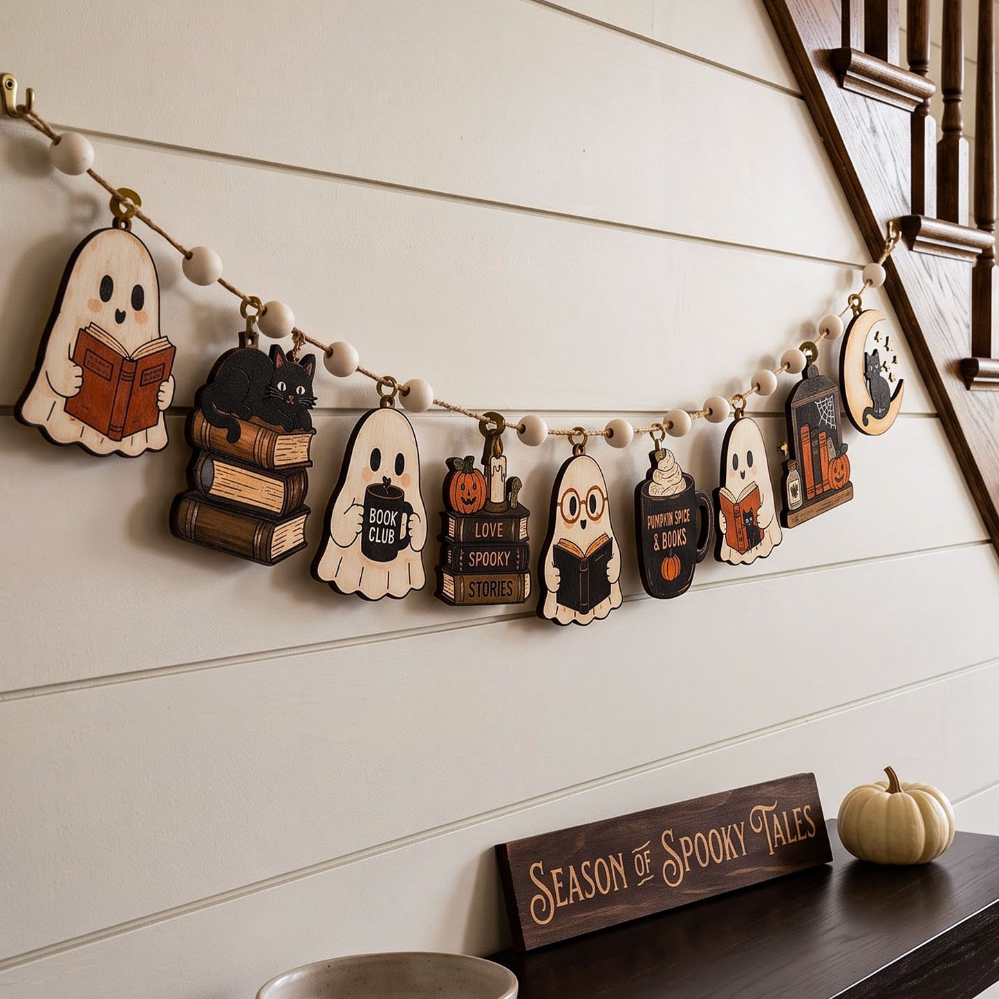 A Halloween-themed wooden bead garland with book and ghost cutouts hangs on a white paneled wall above a mantel. The garland includes shapes like a ghost reading, a black cat on books, a pumpkin with 'I Love Spooky Stories,' a wizard, a cupcake with a book, and a haunted house.