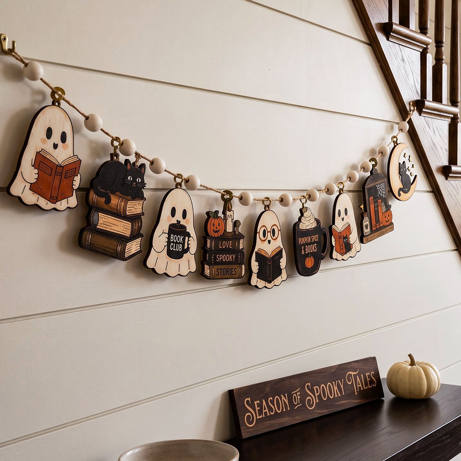 A Halloween-themed wooden bead garland with book and ghost cutouts hangs on a white paneled wall above a mantel. The garland includes shapes like a ghost reading, a black cat on books, a pumpkin with 'I Love Spooky Stories,' a wizard, a cupcake with a book, and a haunted house.