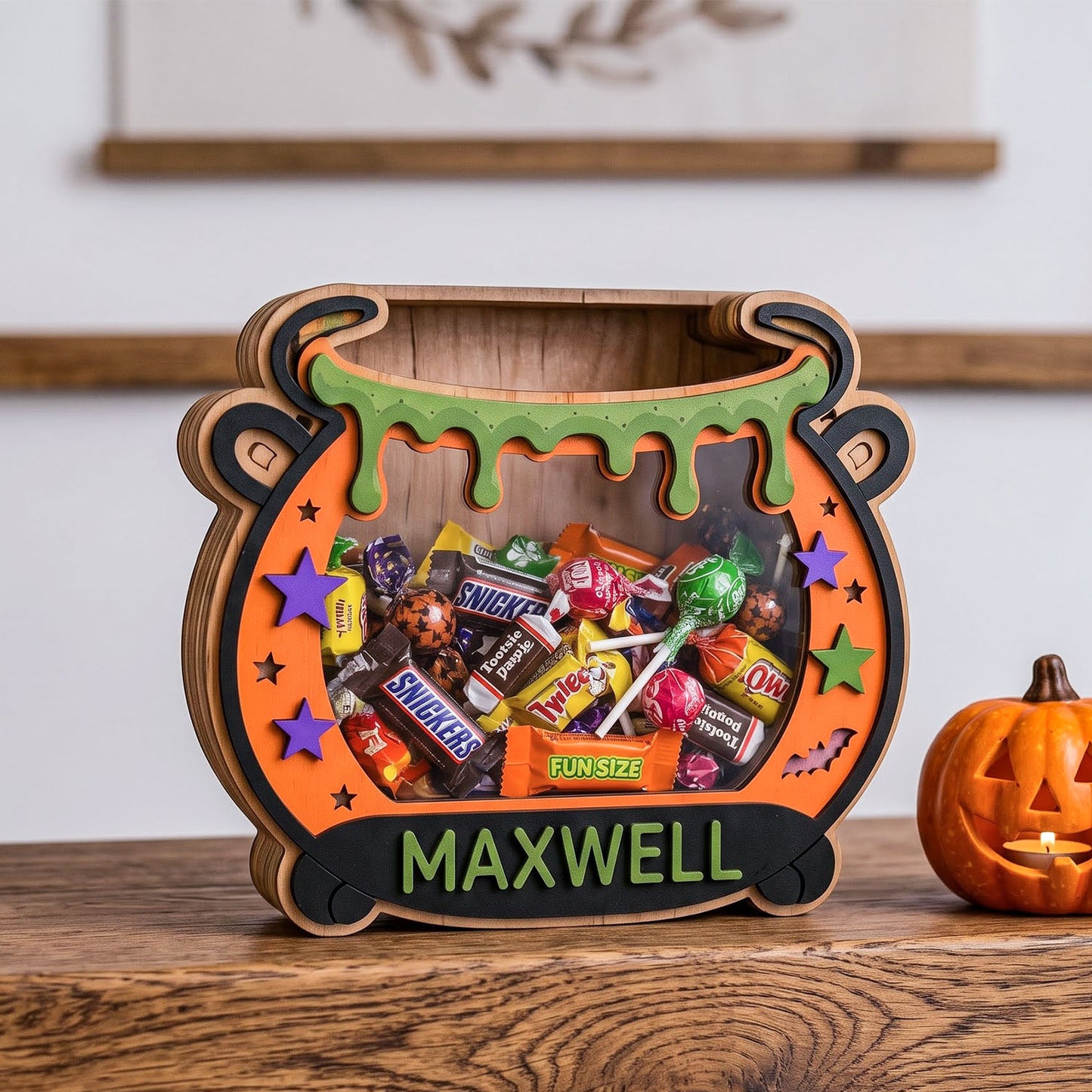 A personalized wooden cauldron-shaped candy box filled with assorted Halloween treats, displayed on a wooden table next to a small jack-o'-lantern. The box features a custom name, 'MAXWELL', in green lettering on a black banner at the bottom.