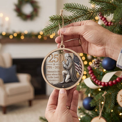 A person holds a round wooden Christmas ornament featuring a kneeling figure and the text '250 1776–2026 Blessed is the nation whose God is the Lord. PSAOM 28:2' in front of a decorated Christmas tree in a cozy living room.