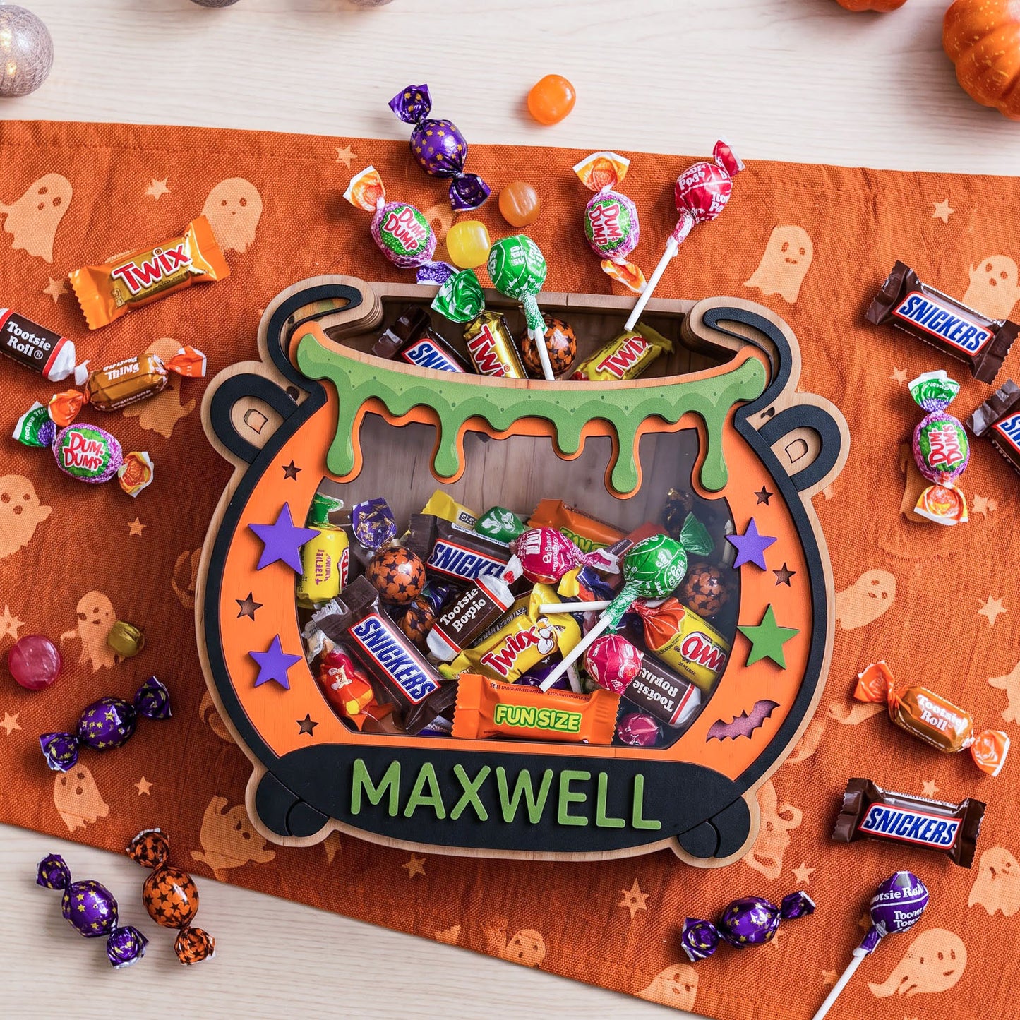 A Halloween-themed wooden cauldron-shaped candy display filled with assorted lollipops and wrapped candies, placed on an orange ghost-patterned runner with scattered Snickers Twix, Kit Kat, and other candy pieces around it.