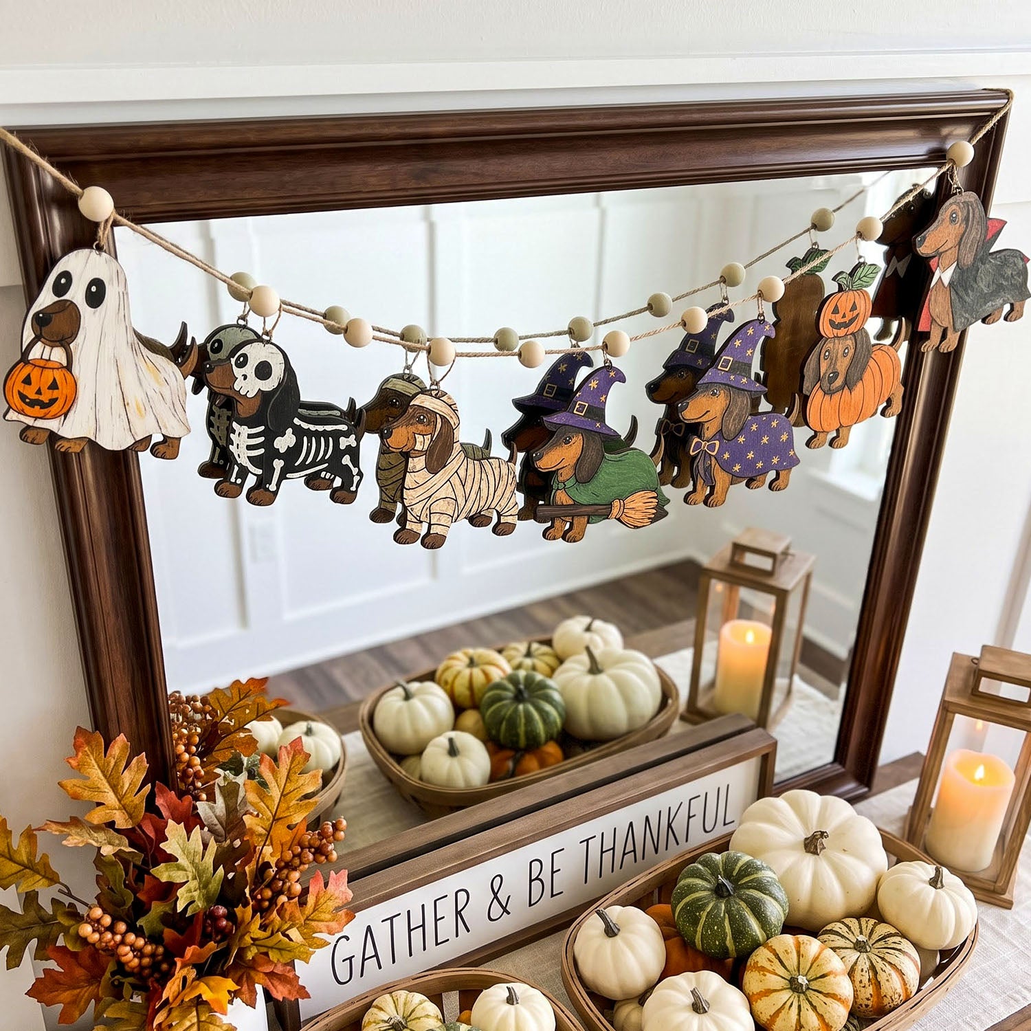 A wooden bead garland featuring cute dog Halloween costumes hangs across a dark wood mirror in a decorated fall entryway. The dog designs include a ghost, skeleton, mummy, witch, pumpkin, and turkey, with some beads showing painted dog faces.