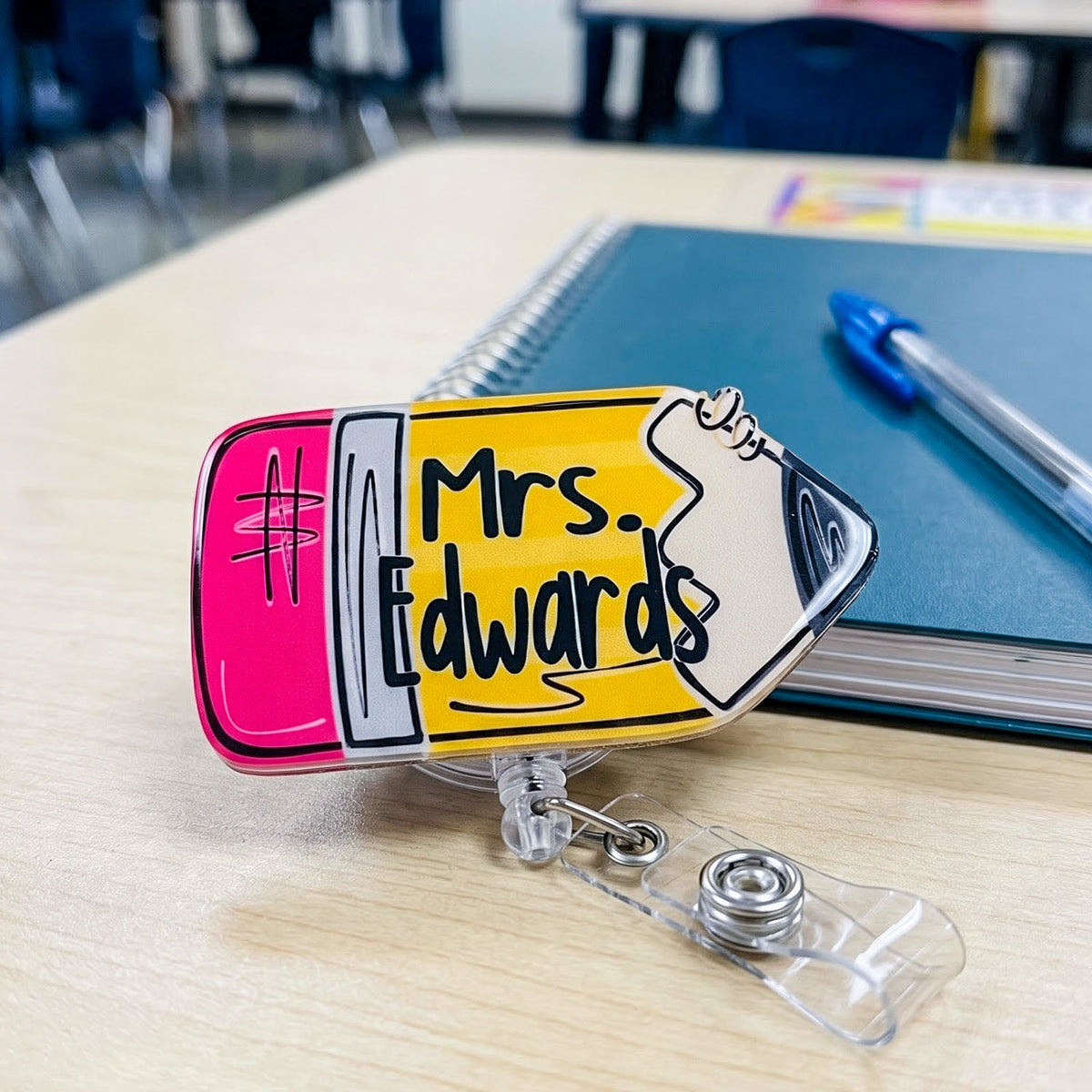 A novelty classroom badge reel shaped like a yellow pencil eraser with the text 'Mrs. Edwards' and a pink eraser head, shown on a light wood desk with a notebook and pen in the background.