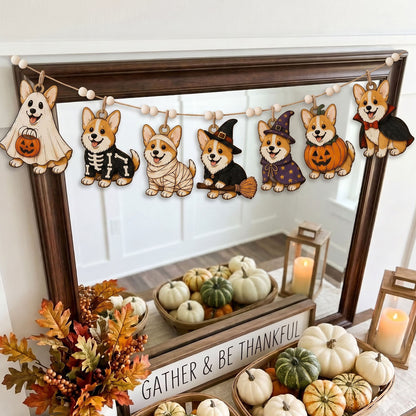 A Halloween-themed wooden dog garland with seven cutout dogs dressed in ghost, skeleton, bandage, witch, wizard, pumpkin, and vampire costumes, hanging across a dark wood mirror in a decorated fall display with pumpkins and candles.
