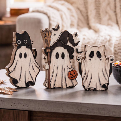 Three Halloween-themed wooden ghost decorations displayed on a tabletop in a cozy living room setting. The set includes a black cat on a white ghost, a witch holding a broomstick with a jack-o'-lantern, and a white ghost with black cat-like features.
