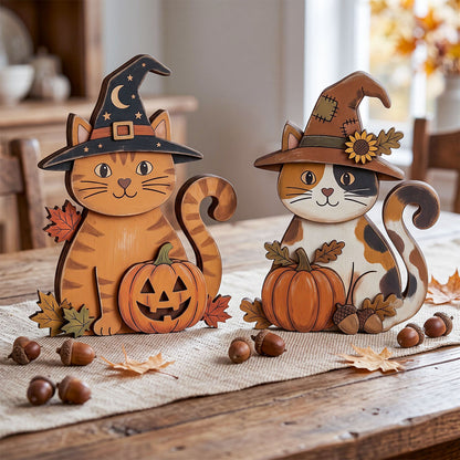 Two decorative wooden cat figurines dressed as witches sit on a rustic wooden table, each holding a carved pumpkin. One cat is orange tabby with a black witch hat, and the other is white and brown with a brown witch hat decorated with a sunflower.