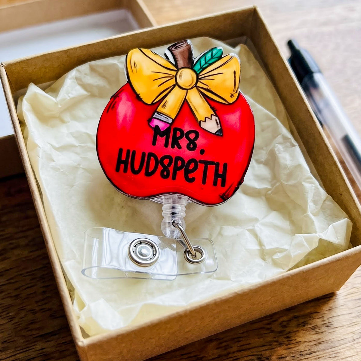 A red apple-shaped badge reel with a yellow bow, pencils, and the text 'MR8. HUDSPETH' displayed inside a brown kraft box on tissue paper.