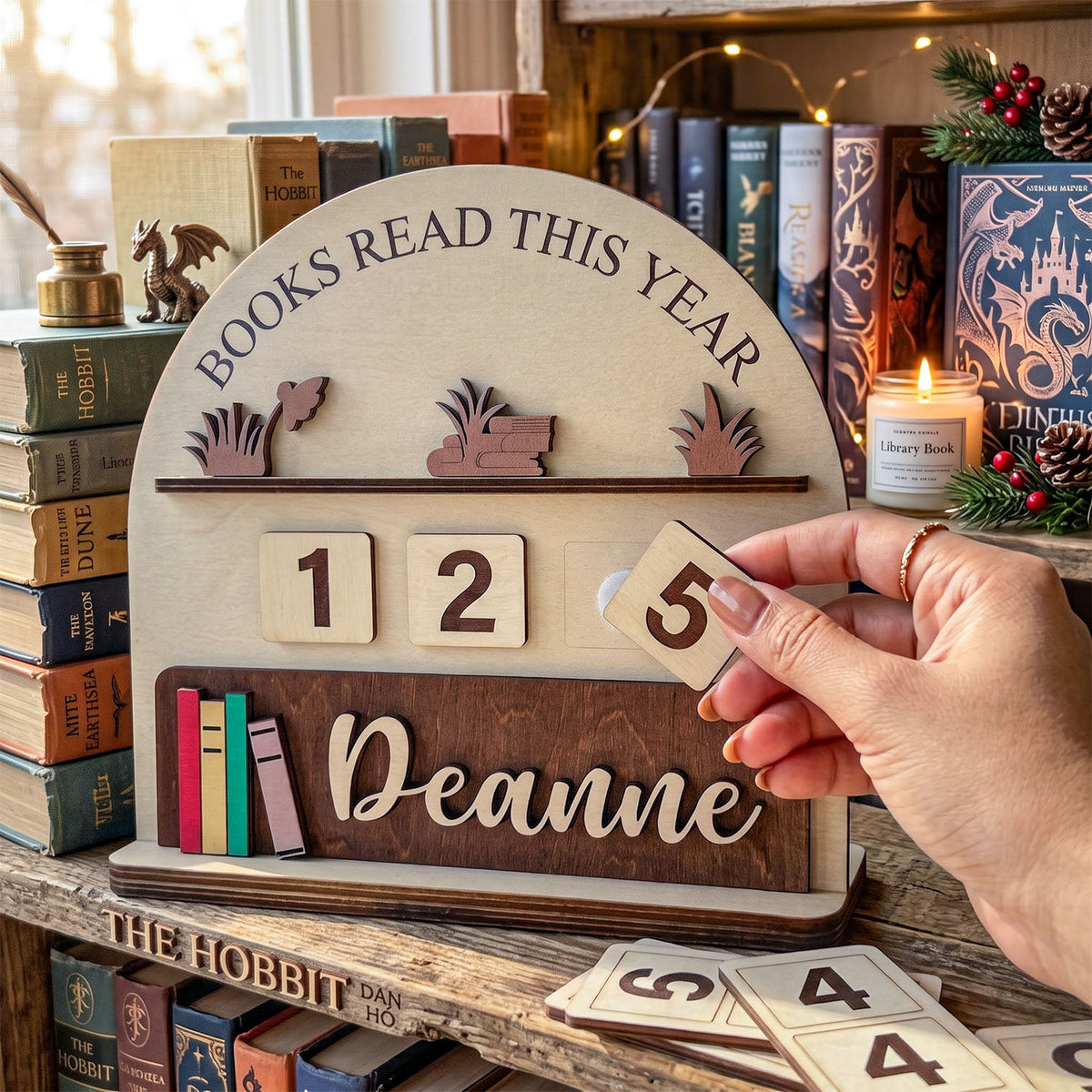 Personalized wooden 'Books Read This Year' shelf sign with sliding number tiles and a nameplate, shown on a bookshelf with a hand holding one tile.