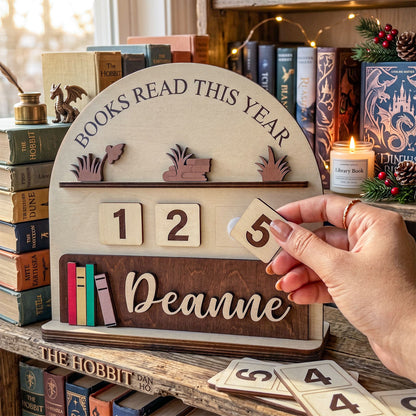 Personalized wooden 'Books Read This Year' shelf sign with sliding number tiles and a nameplate, shown on a bookshelf with a hand holding one tile.