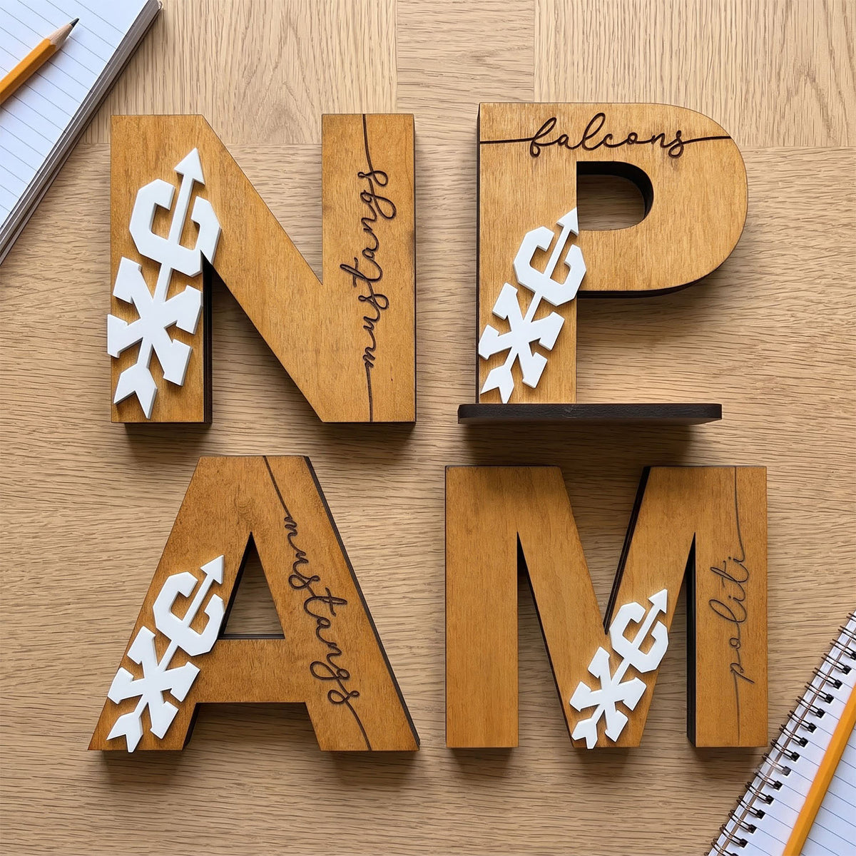 Four wooden block letters arranged to spell 'N P A M' on a light wood tabletop, each engraved with a different script word ('balcons', 'mustangs', 'mustangs', 'politi') and decorated with white paper snowflake cutouts. A pencil, notebook, and another wooden letter are partially visible at the edges.