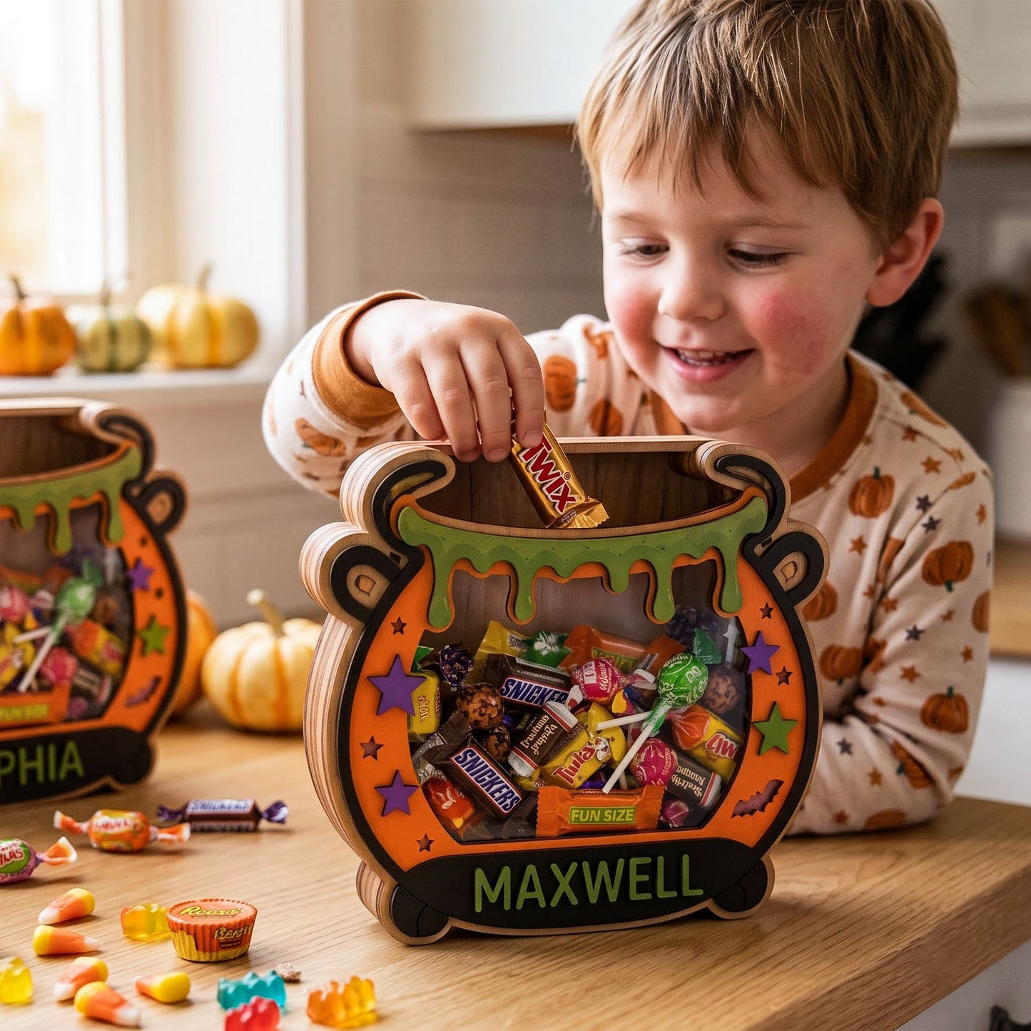 A young boy in a Halloween-themed pajama shirt smiles as he reaches into a personalized wooden cauldron-shaped candy box labeled 'MAXWELL' on a wooden table. The cauldron is decorated with orange, green, and purple Halloween graphics and is filled with various candies.