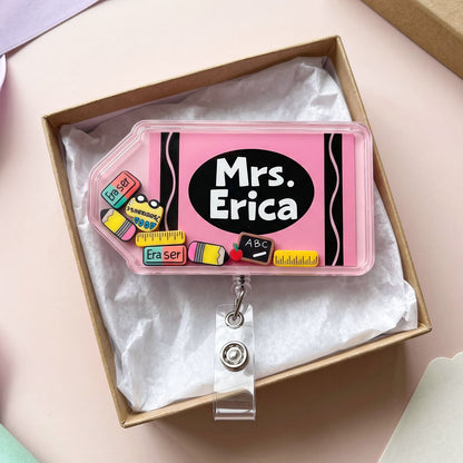 A pink plastic badge reel with a black oval label reading 'Mrs. Erica' and school-themed decorative charms, displayed inside a brown gift box with white tissue paper on a pale pink surface.